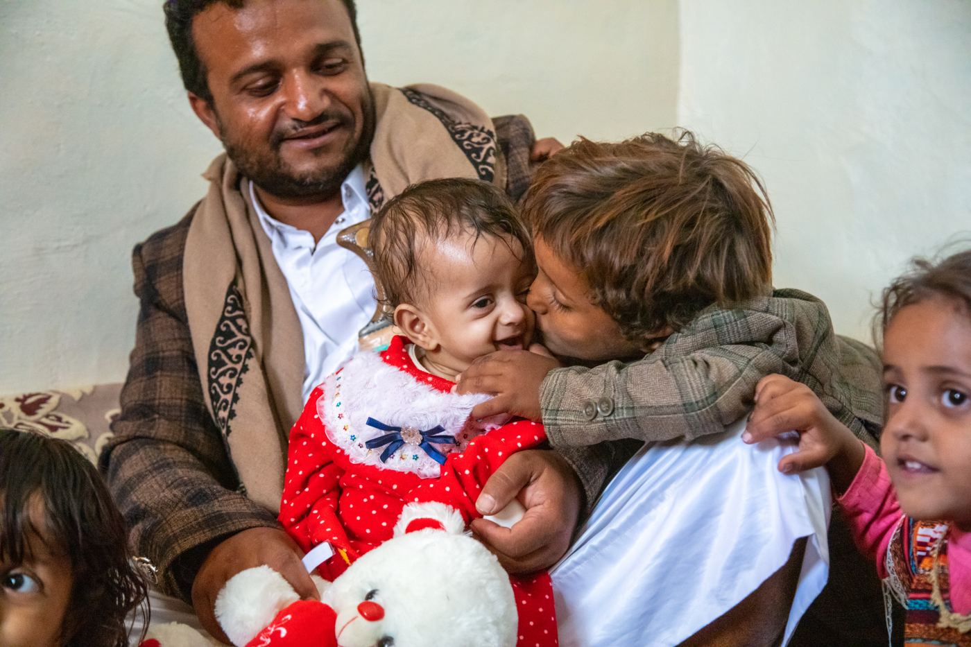 A father sits holding two young children in a therapeutic feeding centre; one child kisses the other on the cheek, Harf Sufyan, Yemen.