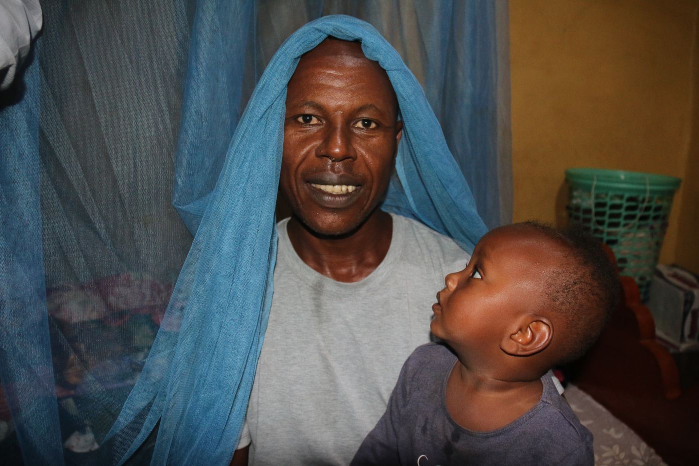 A father sits under a blue insecticide-treated bed net holding his small child, who looks up at him; joint delivery of malaria vaccines and bed nets, Ethiopia.