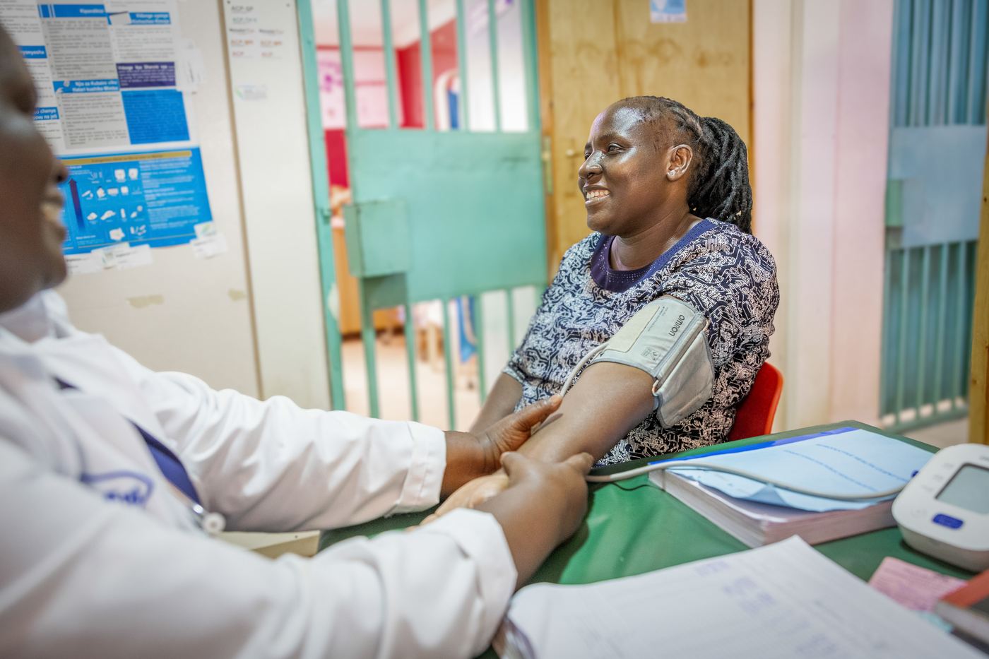 A clinician in a white coat takes the blood pressure of a seated patient during a Women's Integrated Cancer Services (WICS) visit that combines breast and cervical cancer screening with diabetes, hypertension and mental-health checks, Kenya.